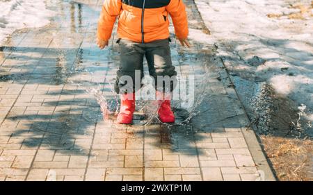 Fröhliche Pfützen : mit jedem Schritt senden die roten Stiefel des Kindes eine Kaskade von Tropfen in die Luft, eine Feier des schmelzenden Schnees an einem sonnigen Tag Stockfoto