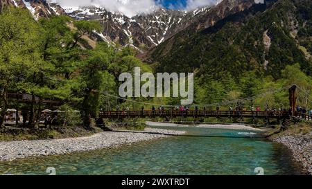 KAMIKOCHI, JAPAN - 24. MAI 2023: Wanderer und Touristen auf Kappabashi mit den schneebedeckten Gipfeln des Kotaka-Gebirges dahinter. Kamikochi, Präfektur Nagano Stockfoto