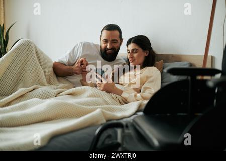 Fröhliche Frau mit Mobilitätseinschränkung, die im Bett liegt, mit Telefon neben ihrem bärtigen, liebevollen Ehemann Stockfoto