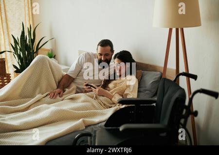 Fröhliche Frau mit Mobilitätseinschränkung, die im Bett liegt, mit Telefon neben ihrem bärtigen, liebevollen Ehemann Stockfoto
