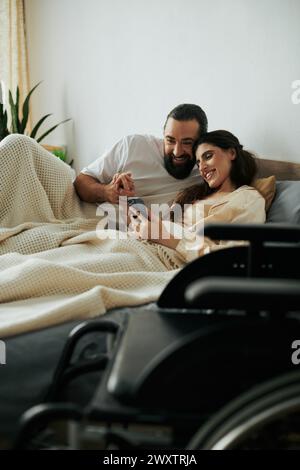 Fröhliche Frau mit Mobilitätseinschränkung, die im Bett liegt, mit Telefon neben ihrem bärtigen, liebevollen Ehemann Stockfoto