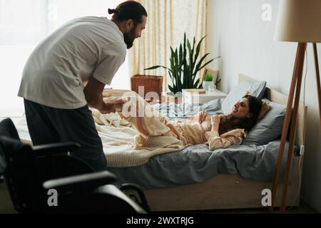 Bärtiger, gutaussehender Mann, der seiner behinderten, fröhlichen Frau hilft, die Muskeln zu dehnen, während sie auf dem Bett liegt Stockfoto