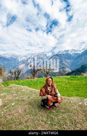 Eine einheimische alte Frau, die einen Schal trägt, hockt auf einem Feld im Dorf am Naddi Aussichtspunkt, berühmt für den Blick auf die riesige Himalaya Dhauladhar Range Stockfoto