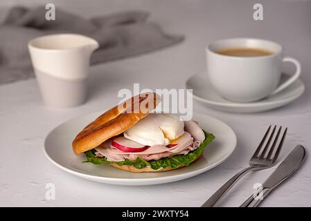 Lebensmittelfotografie von Bagel mit pochiertem Ei, Rettich, Speck, Schinken, Kaffee, Cappuccino, Latte; Romaine-Salat; Sandwich; morgens; in Scheiben geschnitten; Frühstück; brun Stockfoto