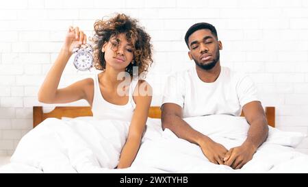 Verschlafenes afroamerikanisches Paar, das mit Uhr im Bett sitzt Stockfoto