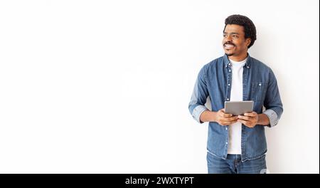 Entspannter schwarzer Mann in Denim-Kleidung, der ein digitales Tablet hält und zur Seite blickt Stockfoto
