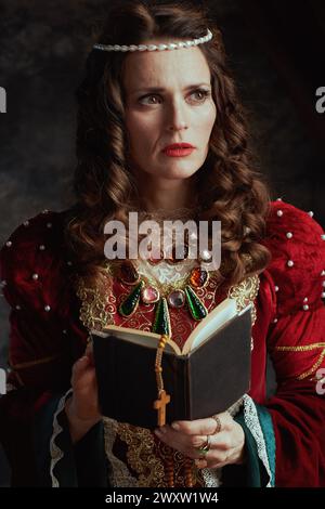 Nachdenkliche mittelalterliche Königin in rotem Kleid mit Buch und Rosenkranz auf dunkelgrauem Hintergrund. Stockfoto