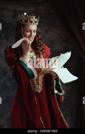 Nachdenkliche mittelalterliche Königin in rotem Kleid mit Pergament und Krone auf dunkelgrauem Hintergrund. Stockfoto