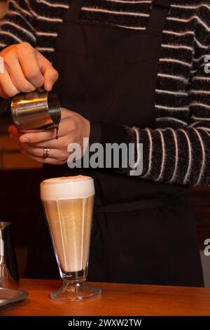 Ein erfahrener Barista in einem stilvollen gestreiften Hemd und einer schwarzen Schürze gießt Sirup in ein Glas aromatischen Kaffee auf einem Holztisch in sanfter Beleuchtung und schafft so etwas Stockfoto