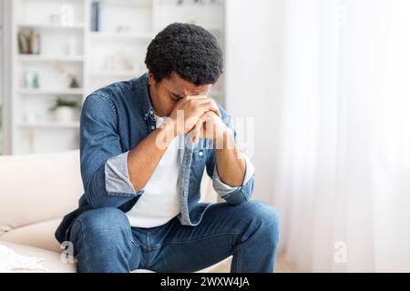 Der junge Schwarze, der auf der Couch sitzt und die Hände das Gesicht bedeckt Stockfoto