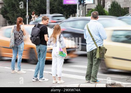 Menschen mit Taschen stehen auf dem Bürgersteig und warten darauf, eine belebte Straße zu überqueren. Die Rückansicht mit dem in Bewegung befindlichen Verkehr verschwimmt an einem Frühlingstag Stockfoto