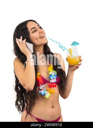 Die junge Lateinfrau, die einen Bikini trägt, spricht auf ihrem Smartphone, während sie einen Sommercocktail in der anderen Hand hält. Sie wirkt entspannt und engagiert, en Stockfoto