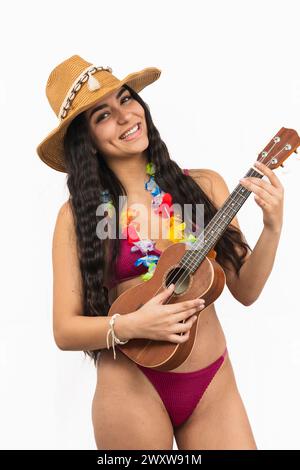 Vertikale Portrait Nahaufnahme lächelnde Latina Frau im Bikini wird gesehen, wie sie die Ukulele spielt. Sie trägt eine Blumenkette und einen Strandhut und strahlt Jo aus Stockfoto