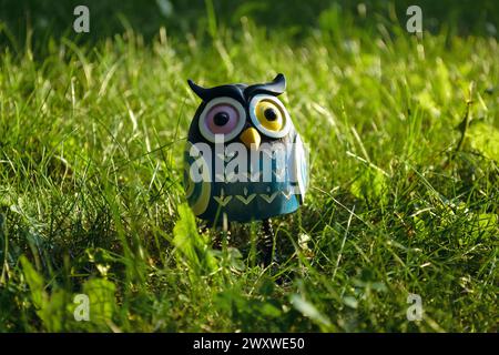 Ein Spielzeug einer überraschten blauen Eule mit mehrfarbigen Augen in Nahaufnahme steht mitten im grünen Gras im Licht der Abendsonne Stockfoto