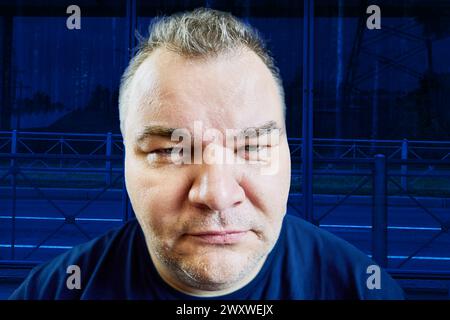 Vorgetäuschte Strenge auf dem Gesicht eines übergewichtigen europäischen Mannes, 47 Jahre alt, horizontales Nahporträt auf blauem Hintergrund, groteske Verzerrung von weit Stockfoto