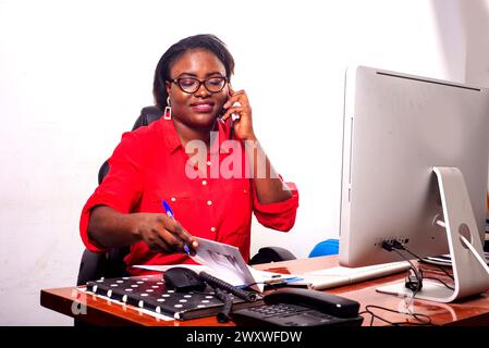 Eine charmante Geschäftsfrau in Brille, die am Schreibtisch vor dem Computer sitzt und am Handy spricht, während sie einen Notizblock überprüft, lächelt. Stockfoto