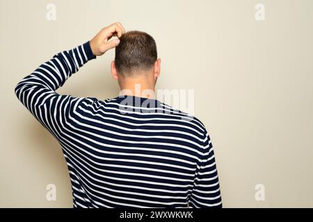 Der junge hispanische Mann kratzt sich den Kopf mit dem Rücken zur Kamera, er hat Zweifel daran, wie man ein Problem lösen kann. Isoliert auf beigefarbenem Studiohintergrund. Stockfoto