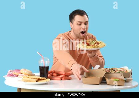Schockierter junger Mann am Tisch voller ungesunder Nahrung auf blauem Hintergrund. Das Konzept des Überessens Stockfoto