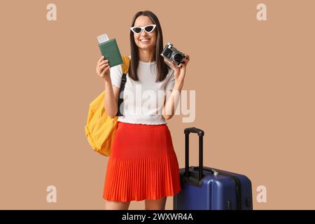 Junge glückliche Frau in Sonnenbrille mit Pass, Fotokamera und Koffer auf beigefarbenem Hintergrund Stockfoto