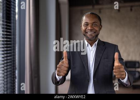 Fröhlicher afroamerikanischer Geschäftsmann im Anzug zeigt den doppelten Daumen hoch und symbolisiert Anerkennung und Erfolg in einer Büroumgebung. Stockfoto