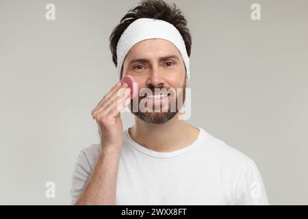 Mann mit Stirnband, der sein Gesicht mit Schwamm auf hellgrauem Hintergrund wäscht Stockfoto