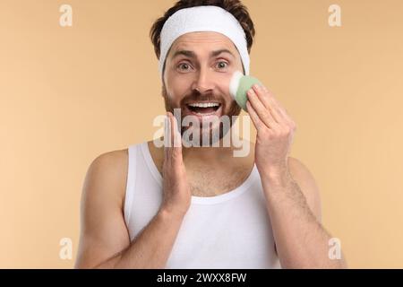 Mann mit Stirnband, der sein Gesicht mit einem Schwamm auf beigem Hintergrund wäscht Stockfoto