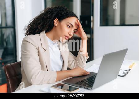 Traurige brasilianische oder hispanische Geschäftsfrau mit lockhaarigem Haar, Corporate Financial Manager, sitzt in einem modernen Büro, schaut traurig auf einen Laptop-Bildschirm, liest schlechte Nachrichten, erhält schlechte Finanzberichte Stockfoto