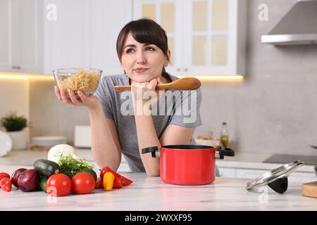 Nachdenkliche Hausfrau mit Löffel und roher Pasta am weißen Marmortisch in der Küche. Kochprozess Stockfoto