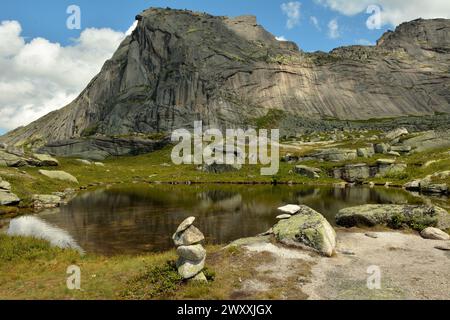Rituelle Steinpyramiden am Ufer eines kleinen Sees mit einer Reflexion eines hohen Berges unter einem wolkigen Sommerhimmel. Naturpark Ergaki, Krasnoya Stockfoto