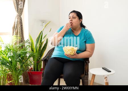 Übergewichtige, fette indianerin sitzt auf einem Stuhl und isst zu Hause Popcorn. Große Frauen genießen Snacks. Ungesunde Ernährung. Stockfoto