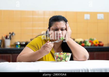 Traurige übergewichtige, fette indianerin, die Gabel hält und Gemüsesalat isst, während sie auf einem Tisch in der Küche sitzt. Plus-Größe weiblich. Diätekampf im Geheimen Stockfoto