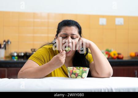 Traurige übergewichtige, fette indianerin, die Gabel hält und Gemüsesalat isst, während sie auf einem Tisch in der Küche sitzt. Plus-Größe weiblich. Diätekampf im Geheimen Stockfoto