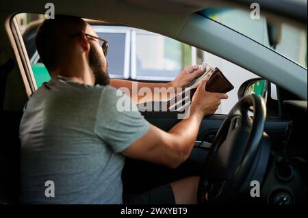 Junger Mann, der Benzin in bar bezahlt. Mann, der Geld aus der Brieftasche holt, um Kraftstoff zu bezahlen. Konzept für hohe Kraftstoffpreise. Stockfoto