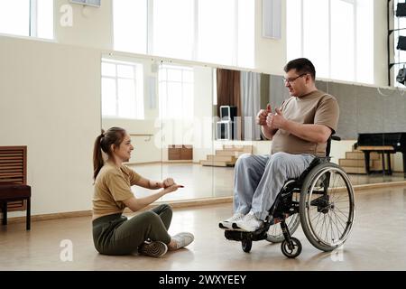 Langer Schuss einer jungen Frau, die vor reifem Mann im Rollstuhl auf dem Boden sitzt und vor dem Tanztraining Aufwärmübungen macht Stockfoto