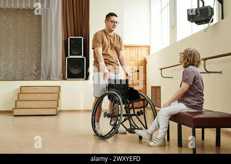 Lange Aufnahme einer jungen Frau mit Behinderung, die auf einer Bank im Tanzstudio sitzt und mit einem jungen Mann spricht Stockfoto
