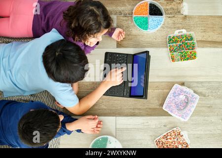 Von oben sehen Kinder, die mit hama-Perlen spielen und mit einem Laptop arbeiten, was eine Mischung aus traditionellem Kunsthandwerk und digitalem Lernen veranschaulicht Stockfoto