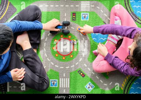 Blick von oben auf einen Jungen und ein Mädchen, der mit Spielzeugautos auf einer lebendigen Spielmatte mit Straßenmotiven spielt, wobei der Fokus auf ihre Hände und die Spielzeuge gerichtet ist. Stockfoto