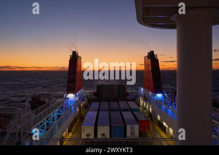 Frachtschiff in der Dämmerung mit beleuchteten Decks und Containern unter Sonnenuntergang gefangen Stockfoto