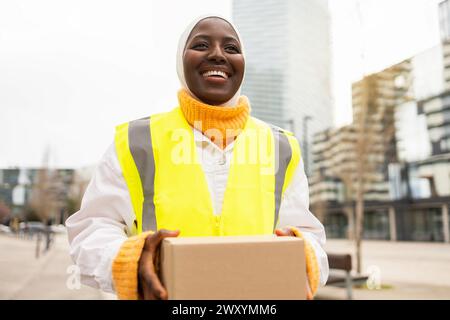Eine fröhliche afroamerikanische Zustellerin in einer gut sichtbaren Weste, die ein Paket in einer städtischen Umgebung mit Gebäuden im Hintergrund hält Stockfoto