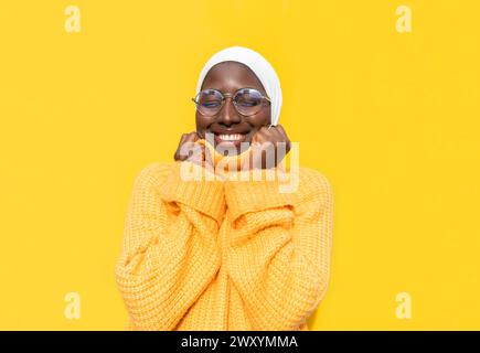 Eine fröhliche Afroamerikanerin mit geschlossenen Augen, die einen weißen Hijab und einen bequemen gelben Pullover vor einem hellgelben Hintergrund trägt und Glück ausstrahlt Stockfoto