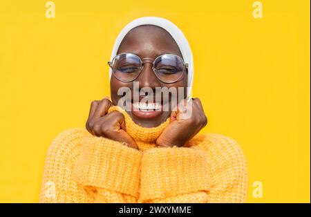 Eine fröhliche Afroamerikanerin mit geschlossenen Augen, die einen weißen Hijab und einen bequemen gelben Pullover vor einem hellgelben Hintergrund trägt und Glück ausstrahlt Stockfoto