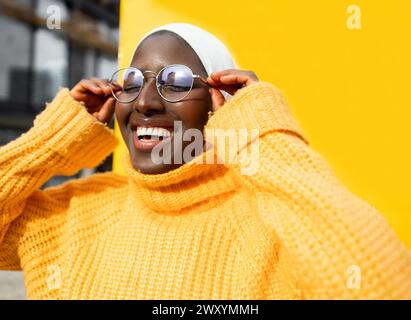 Lächelnde schwarze Frau in gelbem Pullover, die Brille vor einem leuchtend gelben Hintergrund justiert und in die Kamera blickt. Stockfoto