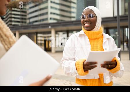Zwei afroamerikanische Frauen führen auf einer Straße eine freundliche Unterhaltung, die Wärme und Kameradschaft vor einem urbanen Hintergrund ausstrahlt. Stockfoto