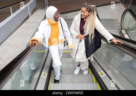 Zwei fröhliche afroamerikanische Frauen, die sich in einer städtischen Umgebung auf eine Rolltreppe begeben. Stockfoto