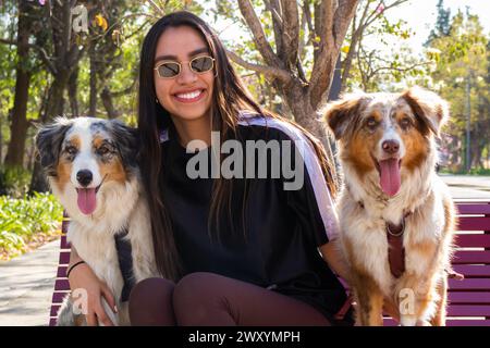 Eine fröhliche junge Dame in Sonnenbrille sitzt auf einer Parkbank mit zwei glücklichen australischen Schäferhunden, die einen sonnigen Tag genießen Stockfoto