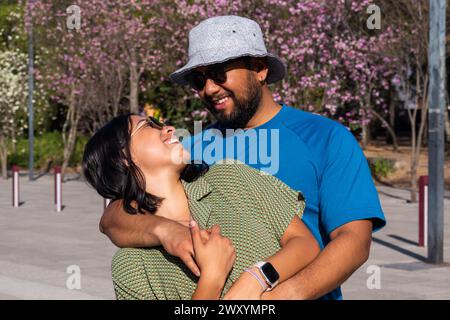 Ein fröhliches Paar genießt einen sonnigen Tag im Freien und teilt eine warme Umarmung mit blühenden rosa Bäumen im Hintergrund Stockfoto