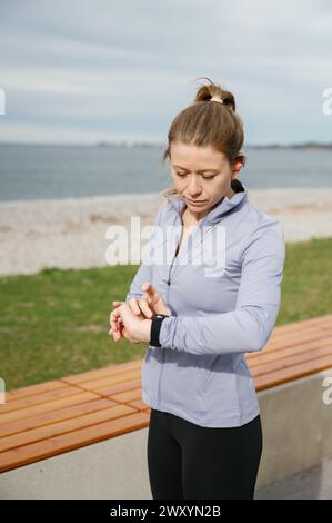 Eine fokussierte Frau passt ihre Smartwatch nach einem Workout am Meer an, mit einem bewölkten Himmel im Hintergrund Stockfoto