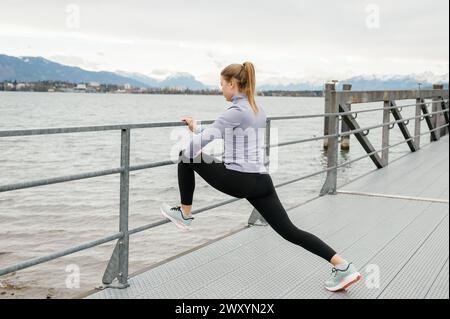 Eine fokussierte Frau streckt die Beine an einem Geländer entlang einer malerischen Uferpromenade mit Bergen in der Ferne Stockfoto