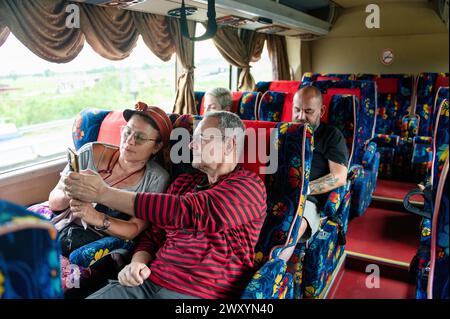 Ein älteres Paar sitzt eng zusammen und teilt sich ein Smartphone, während es in einem farbenfrohen Reisebus unterwegs ist, während ein anderer Passagier im Hintergrund schläft Stockfoto