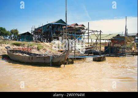 Ein traditionelles Holzboot, das entlang eines Flusses mit Pfahlhäusern und Gerüsten in einem schwimmenden Dorf unter einem klaren blauen Himmel vertäut ist Stockfoto